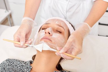 Güzellik uzmanı, güzellik salonunda estetik krem maskesini cilt tazeleme, nemlendirici kozmetik işlemleri için kadın yüzüne uyguluyor. Eldivenli kozmetik uzmanı iki fırçayla kozmetik krem maskesi takıyor.