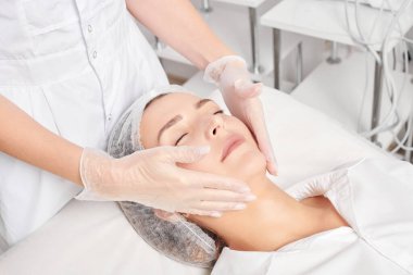 Kozmetik uzmanı, güzellik salonundaki gençleştirme için nemlendirici krem sürdükten sonra cildine masaj yapıyor. Eldivenli güzellik uzmanı elleri rahatlamak için kadın yüzüne masaj yapıyor.