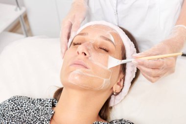 Kozmetik uzmanı, güzellik salonundaki kozmetik bakım ürünlerini nemlendirmek için kadın yüzüne estetik krem takıyor. Eldivenli güzellik uzmanı fırçayla kozmetik krem maskesi takıyor.