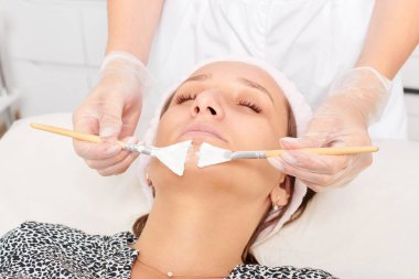 Kozmetik uzmanı, güzellik salonunda cildini yenilemek için kadın yüzüne kozmetik krem maskesi uyguluyor. Eldivenli güzellik uzmanı iki fırçalı kozmetik krem maskesi takıyor.