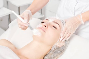 Güzellik uzmanı, güzellik salonundaki yaşlanma karşıtı kozmetik işlemler için cildini yenilemek için kadın yüzüne kozmetik krem maskesi sürüyor. Eldivenleri teslim eden kozmetik uzmanı maske takmak için kozmetik fırça tutuyor.