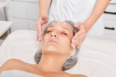 Güzellik uzmanları, güzellik salonlarında gençleştirme için nemlendirici krem maskeyi kadın yüzüne sürerler, yaşlanma karşıtı kozmetik işlemler yaparlar. Kozmetik uzmanı kadın derisi için yüz kremi uyguluyor.