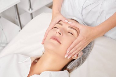 Güzellik uzmanı, güzellik salonunda gençleştirme için nemlendirici krem sürdükten sonra cildine masaj yapıyor, yaşlanma önleyici kozmetik prosedürler uyguluyor. Kozmetik uzmanı rahatlamak için kadın yüzüne masaj yapıyor.