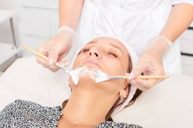 Güzellik uzmanı, güzellik salonunda estetik krem maskesini cilt tazeleme, nemlendirici kozmetik işlemleri için kadın yüzüne uyguluyor. Eldivenli kozmetik uzmanı iki fırçayla kozmetik krem maskesi takıyor.