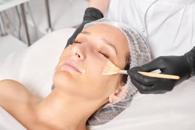 Güzellik uzmanı kadın yüzüne nemlendirici yüz derisi ve güzellik salonundaki yaşlanma karşıtı kozmetik prosedürleri için bal maskesi takıyor. Siyah eldivenli kozmetik uzmanı bal maskesi takmak için kozmetik fırça tutuyor.