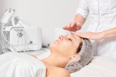 Güzellik salonunda kozmetik uzmanı cildine nemlendirici krem maskeyi sürüp gençleştirme, yaşlanmayı önleyici kozmetik prosedür uyguluyor. Güzellik uzmanı elleri kadın derisi için yüz kremi sürüyor.