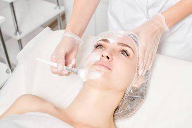 Güzellik uzmanı, güzellik salonlarında cilt bakımı için kadın yüzüne vitamin kremi maskesi, yaşlanma karşıtı kozmetik işlemler uyguluyor. Eldivenli kozmetik uzmanı maske takmak için kozmetik fırça tutuyor