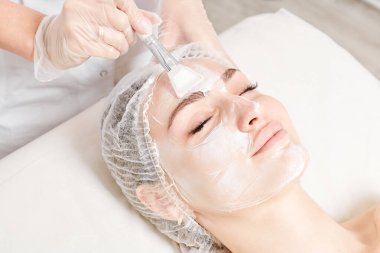 Güzellik uzmanı, güzellik salonlarında cilt bakımı için kadın yüzüne vitamin kremi maskesi, yaşlanma karşıtı kozmetik işlemler uyguluyor. Eldivenli kozmetik uzmanı maske takmak için kozmetik fırça tutuyor