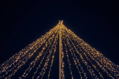 Sarı ışık çelenkleri gece mavi gökyüzü arka planında sütunlardan sarkan teller, sihirli tatil atmosferi. Parlak sarı ışıklı Noel çelenkleri, güzel açık hava dekorasyonu.