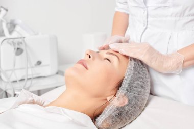 Kozmetik uzmanı, güzellik salonundaki gençleştirme için nemlendirici krem sürdükten sonra cildine masaj yapıyor. Eldivenli güzellik uzmanı elleri rahatlamak için kadın yüzüne masaj yapıyor.