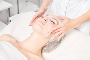 Güzellik uzmanı, güzellik salonundaki cilt bakımı için kadın yüzüne çarşaf maskesi, kırışıklık önleyici kozmetik prosedürler takıyor. Kozmetik uzmanı kadın yüzüne nemlendirici çarşaf maskesi uyguluyor.