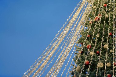 Gece mavi gökyüzü arkaplanındaki sarı çelenkten Noel ağacı ışıkları, kopyalama alanı. Işıl ışıl sarı çelenkleri ve Noel süslemeleri olan parlak Noel ağacı, açık havada tatil atmosferi