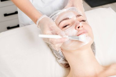 Güzellik uzmanı, güzellik salonundaki yaşlanma karşıtı kozmetik işlemler için cildini yenilemek için kadın yüzüne kozmetik krem maskesi sürüyor. Eldivenleri teslim eden kozmetik uzmanı maske takmak için kozmetik fırça tutuyor.