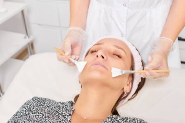 Güzellik uzmanı, güzellik salonunda estetik krem maskesini cilt tazeleme, nemlendirici kozmetik işlemleri için kadın yüzüne uyguluyor. Eldivenli kozmetik uzmanı iki fırçayla kozmetik krem maskesi takıyor.