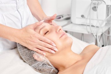 Güzellik salonunda kozmetik uzmanı cildine nemlendirici krem maskeyi sürüp gençleştirme, yaşlanmayı önleyici kozmetik prosedür uyguluyor. Güzellik uzmanı elleri kadın derisi için yüz kremi sürüyor.