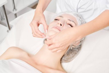 Güzellik uzmanı, güzellik salonundaki cilt bakımı için kadın yüzüne çarşaf maskesi, kırışıklık önleyici kozmetik prosedürler takıyor. Kozmetik uzmanı kadın yüzüne nemlendirici çarşaf maskesi uyguluyor.