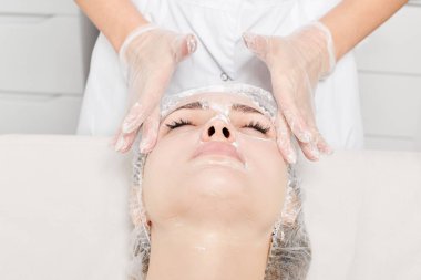 Güzellik uzmanı güzellik salonundaki nemlendirici cilt bakım için kadın yüzüne krem maskesi takıyor. Kozmetik uzmanı kadın yüzüne krem sürerken eldiven verdi.