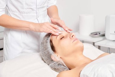 Güzellik uzmanları, güzellik salonlarında gençleştirme için nemlendirici krem maskeyi kadın yüzüne sürerler, yaşlanma karşıtı kozmetik işlemler yaparlar. Kozmetik uzmanı kadın derisi için yüz kremi uyguluyor.