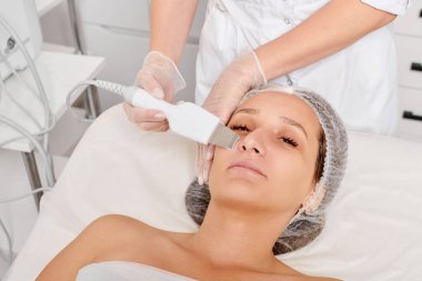 Güzellik uzmanı, güzellik salonundaki yaşlanma karşıtı kozmetik işlemleri ve cilt bakımını yenilemek için yüz ultrasoniği yapıyor. Kozmetik uzmanı yüz bakımını ultrason temizleyicisiyle yapıyor.
