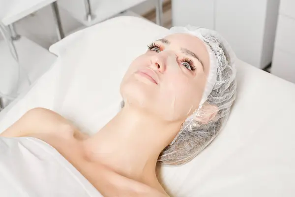 Young woman in sheet mask on face for rehydrate face skin, anti wrinkles cosmetic procedure in beauty spa salon. Beautiful female in moisturizing sheet mask in cosmetologist clinic, skin rejuvenation