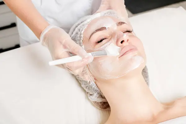 Cosmetologist applies cream mask on woman face for rejuvenation face skin, anti aging cosmetic procedure in beauty spa salon. Beautician hands in gloves holds cosmetic brush and applying cosmetic mask
