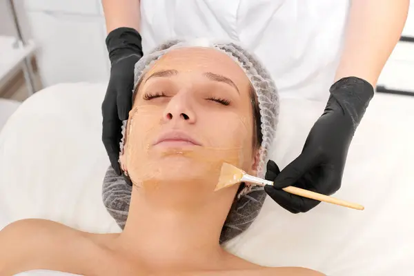 Beautician applies honey mask on woman face for moisturize face skin, anti aging cosmetic procedure in beauty spa salon. Cosmetologist in black gloves holds cosmetic brush for applying honey mask