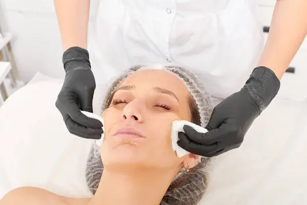 Cosmetologist removing facial cream mask from woman face skin with cosmetic wipes, rejuvenation skincare cosmetic procedure in beauty spa salon. Beautician hands holds napkins for remove face mask