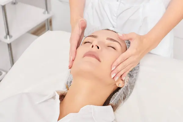 Beautician massages woman face skin after rubbing moisturizing cream for rejuvenation, anti aging cosmetic procedure in beauty spa salon. Cosmetologist hands massages female face for relax