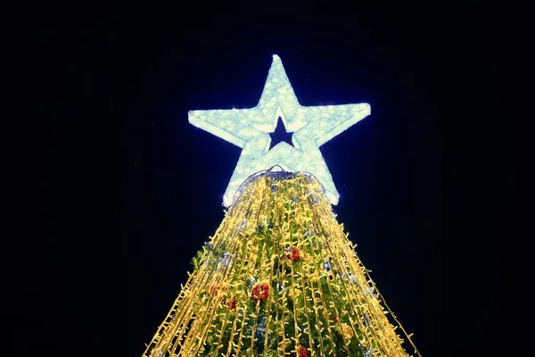 Christmas tree with big white star topper decorated yellow garlands and colorful decorative bulbs on night blue sky background, outdoor holiday atmosphere. Festive Christmas tree with yellow lights