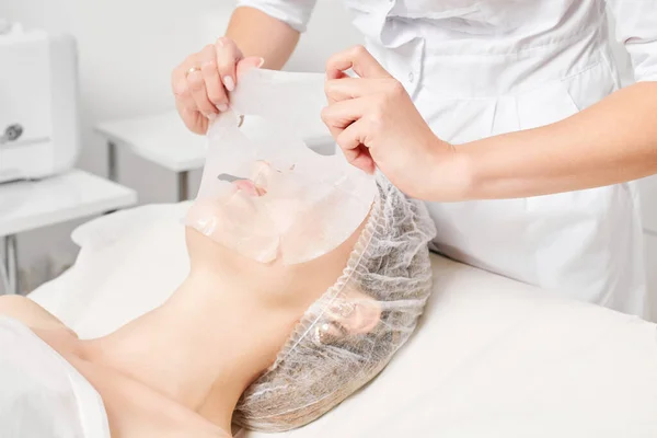 Cosmetologist applying sheet mask on woman face for rehydrate face skin, revitalize cosmetic procedure in beauty spa salon. Beautician applying detox sheet mask on female face