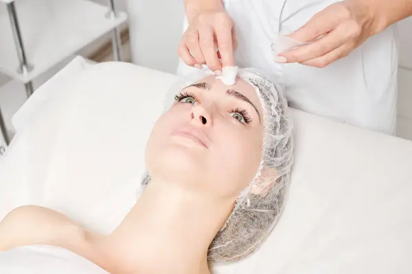 Cosmetologist removing facial cream mask from woman face skin with cosmetic wipes, rejuvenation skincare cosmetic procedure in beauty spa salon. Beautician hands holds napkins for remove face mask