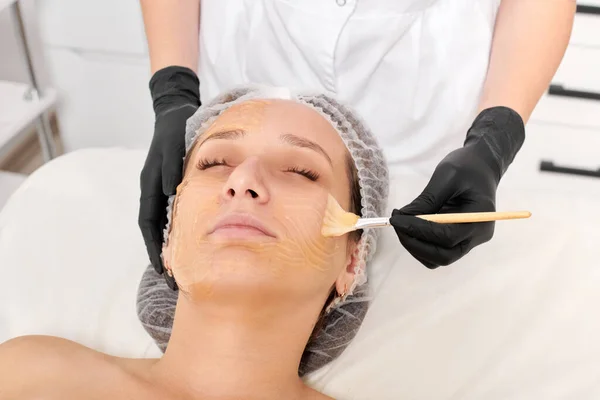 Cosmetologist applies honey mask on woman face for moisturize face skin, anti aging cosmetic procedure in beauty spa salon. Beautician in black gloves holds cosmetic brush for applying honey mask