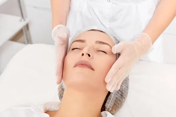 Beautician massages woman face skin after rubbing moisturizing cream for rejuvenation, anti aging cosmetic procedure in beauty spa salon. Cosmetologist hands in gloves massages female face for relax
