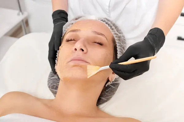 Cosmetologist applies honey mask on woman face for moisturize face skin, anti aging cosmetic procedure in beauty spa salon. Beautician in black gloves holds cosmetic brush for applying honey mask