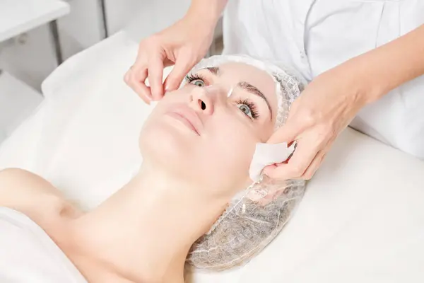 Beautician removing facial cream mask from woman face skin with cosmetic wipes, rejuvenation skincare cosmetic procedure in beauty spa salon. Cosmetologist hands holds napkins for remove face mask