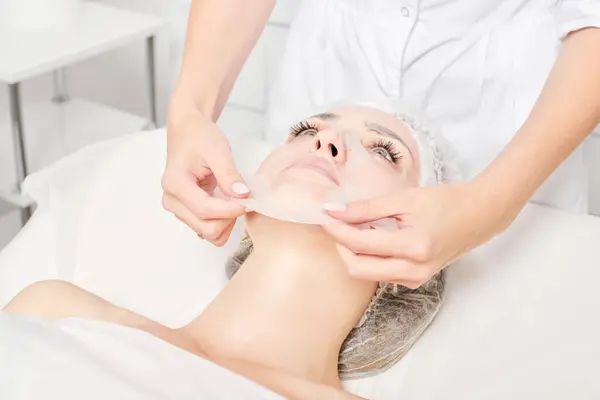 美容师摘除妇女面部的床单面膜 用于再水化面部皮肤 并在美容院进行抗衰老化妆品处理 美容师摘除女性脸部的润肤膜 — 图库照片