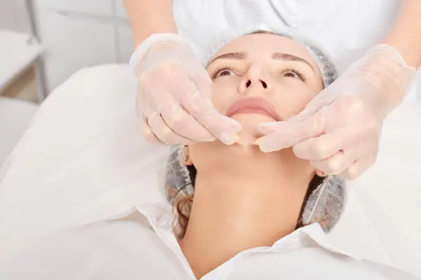 Cosmetologist applies facial oil capsules on woman face for rejuvenation, anti aging cosmetic procedure in beauty spa salon. Beautician hands in gloves holds oil capsules for nourishment