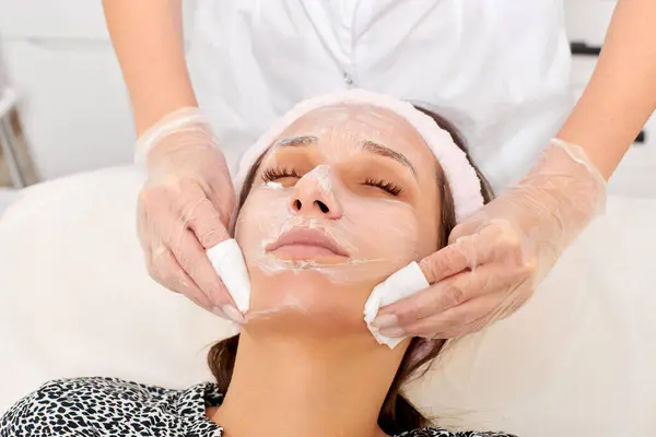 Cosmetologist removing facial cream mask from woman face skin with cosmetic wipes, rejuvenation skincare cosmetic procedure in beauty spa salon. Beautician removing moisturized cream with napkins