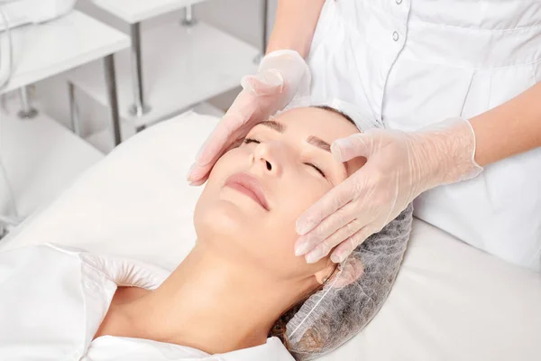 Beautician massages woman face skin after rubbing moisturizing cream for rejuvenation, anti aging cosmetic procedure in beauty spa salon. Cosmetologist hands in gloves massages female face for relax