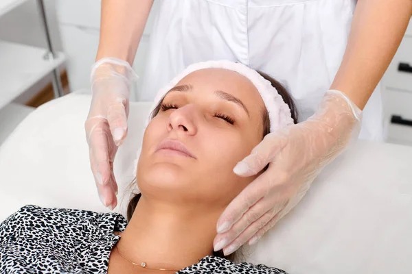 Eldivenli kozmetik uzmanı yenilenme, lenfatik drenaj, güzellik salonundaki krem cilt estetiği için kadın yüz masajı yapıyor. Güzellik uzmanı elleriyle cildini düzleştiriyor.