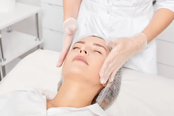 Cosmetologist massages woman face skin after rubbing moisturizing cream for rejuvenation, anti aging cosmetic procedure in beauty spa salon. Beautician hands in gloves massages female face for relax
