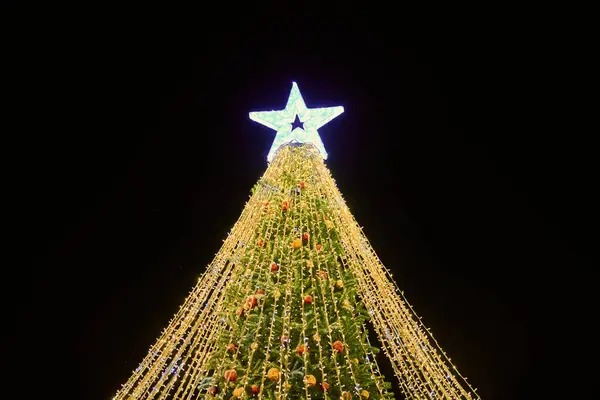 黄色いガーランド 装飾的な球根および夜の青い空の背景 屋外の休日の雰囲気の大きい白い星の上のクリスマス ツリー 明るい黄色のライトガーランドでお祝いのクリスマスツリー ストック画像