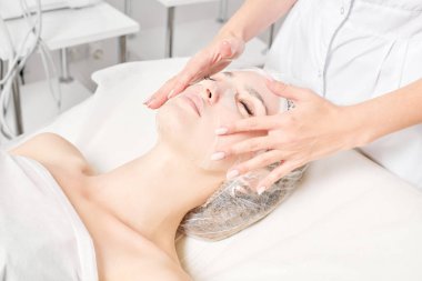 Güzellik uzmanı, güzellik salonundaki cilt bakımı için kadın yüzüne çarşaf maskesi, kırışıklık önleyici kozmetik prosedürler takıyor. Kozmetik uzmanı kadın yüzüne nemlendirici çarşaf maskesi uyguluyor.