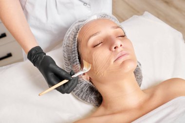 Güzellik uzmanı kadın yüzüne nemlendirici yüz derisi ve güzellik salonundaki yaşlanma karşıtı kozmetik prosedürleri için bal maskesi takıyor. Siyah eldivenli kozmetik uzmanı bal maskesi takmak için kozmetik fırça tutuyor.