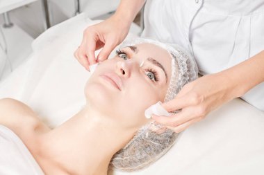 Güzellik uzmanı, güzellik salonundaki yüz kremi maskesini kadın yüzünden kozmetik mendillerle alıyor, cilt yenileme estetik ameliyatı yapıyor. Kozmetik uzmanı yüz maskesini çıkarmak için peçete tutuyor.