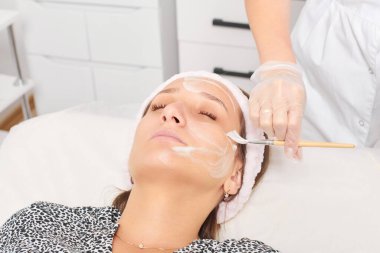 Güzellik uzmanı, güzellik salonundaki kozmetik krem maskesini cilt tazeleme, nemlendirici kozmetik işlemler için kadın yüzüne uyguluyor. Eldivenli kozmetik uzmanı fırçayla kozmetik krem maskesi takıyor.