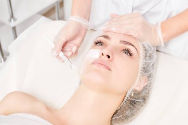 Kozmetik uzmanı kadın yüzüne cilt tazeleme için krem maskesi, güzellik salonundaki yaşlanma karşıtı kozmetik prosedürleri uyguluyor. Eldivenli güzellik uzmanı elleri kozmetik fırça tutuyor ve kozmetik maske takıyor