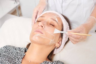 Güzellik uzmanı, güzellik salonundaki kozmetik krem maskesini cilt tazeleme, nemlendirici kozmetik işlemler için kadın yüzüne uyguluyor. Eldivenli kozmetik uzmanı fırçayla kozmetik krem maskesi takıyor.