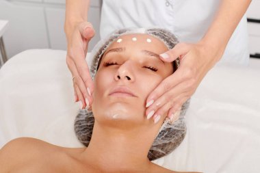 Güzellik salonunda kozmetik uzmanı cildine nemlendirici krem maskeyi sürüp gençleştirme, yaşlanmayı önleyici kozmetik prosedür uyguluyor. Güzellik uzmanı elleri kadın derisi için yüz kremi sürüyor.