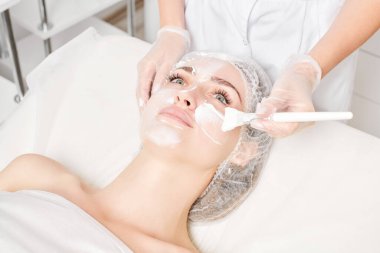 Güzellik uzmanı, güzellik salonundaki yaşlanma karşıtı kozmetik işlemler için cildini yenilemek için kadın yüzüne kozmetik krem maskesi sürüyor. Eldivenleri teslim eden kozmetik uzmanı maske takmak için kozmetik fırça tutuyor.